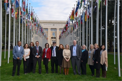 [ai] A group of 12 individuals standing in front of the United Nations building in Geneva, surrounded by flagpoles displaying various national flags. The participants are dressed in professional attire.